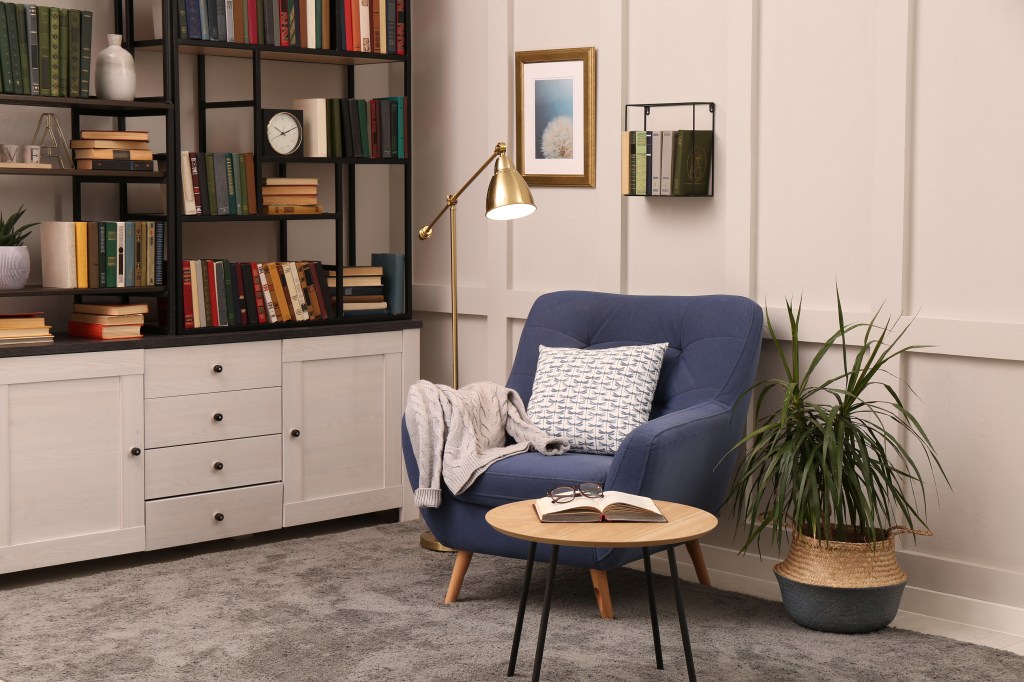 A blue armchair with a blanket and pillow sits beside bookshelves, a reading lamp, and a small table with an open book.