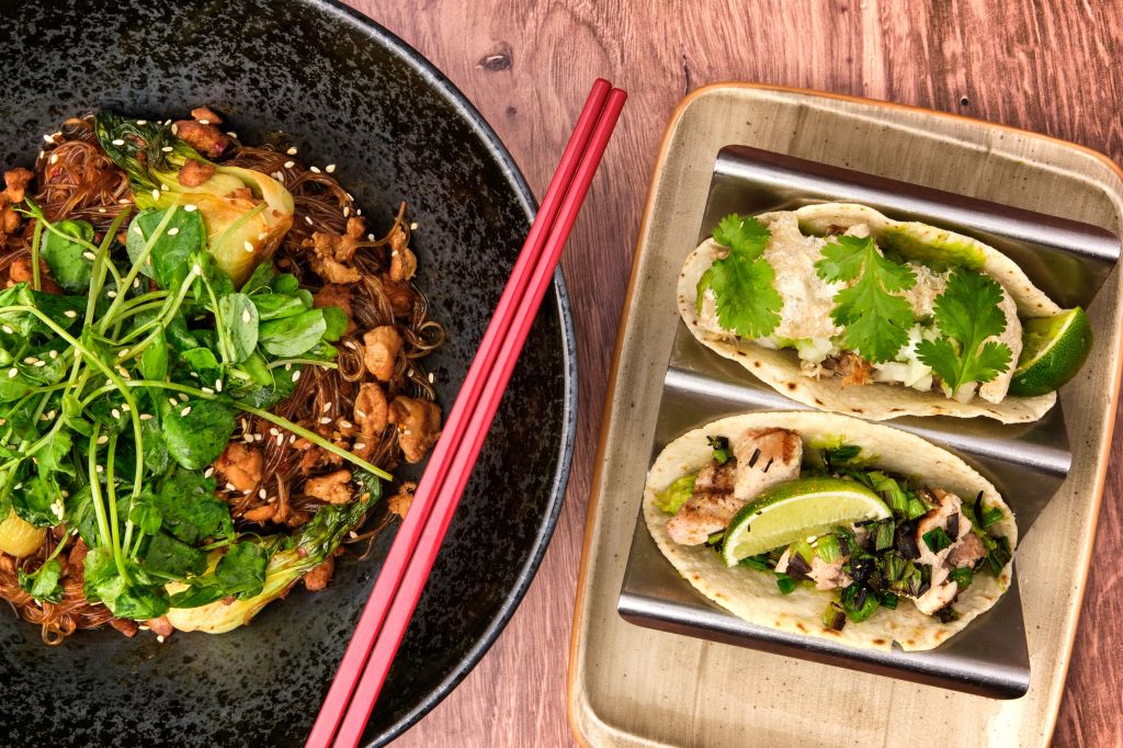 A bowl of noodles topped with greens and sesame seeds beside two tacos garnished with cilantro and lime.