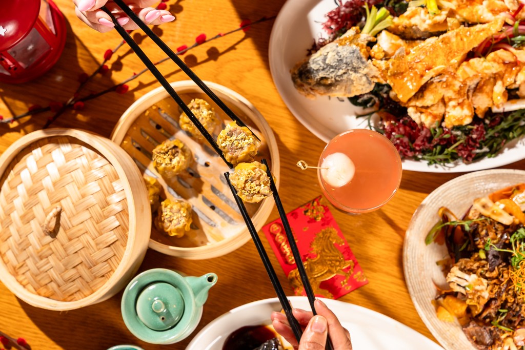 Diners lift scallop and pork siu mai with chopsticks above a bamboo steamer, surrounded by dishes and a pink cocktail.