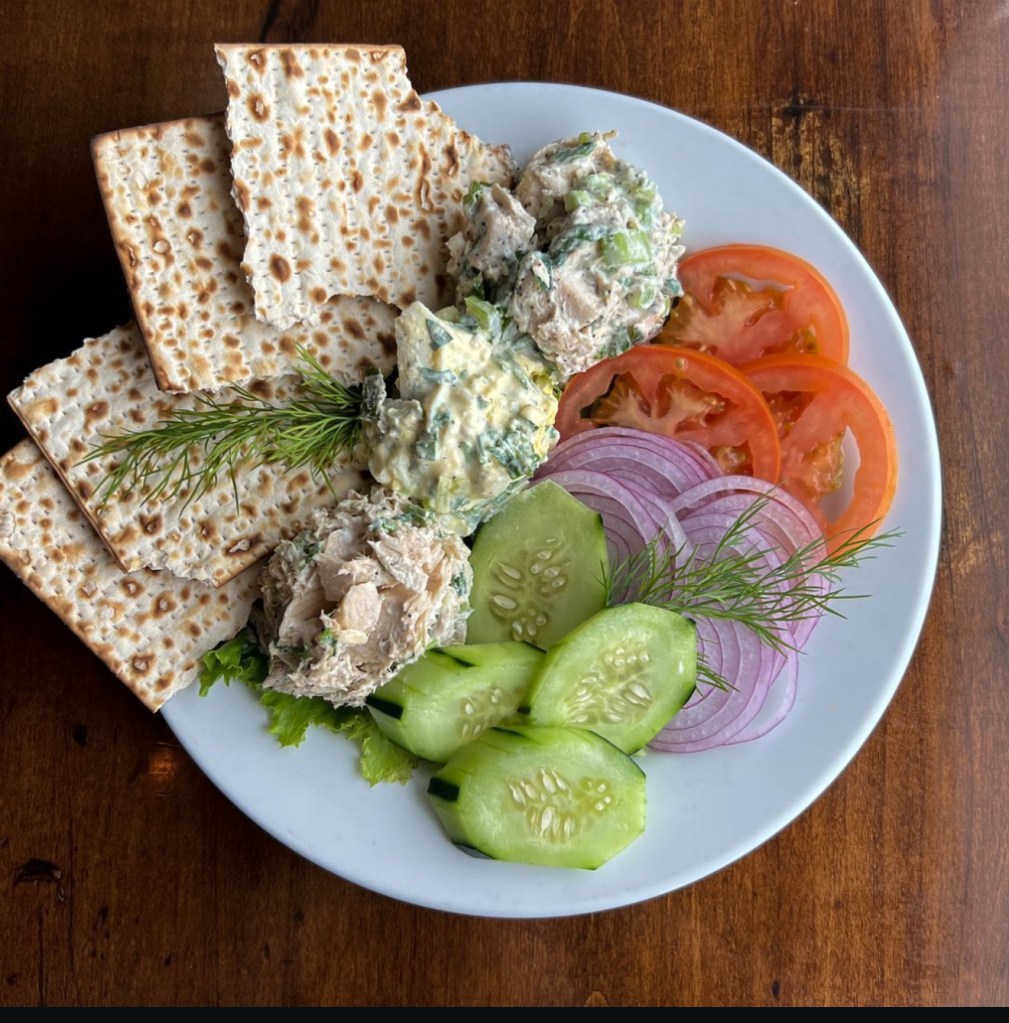 Matzo and scoops of deli salads with sliced tomatoes, cucumbers, red onions, and dill on a white platter.