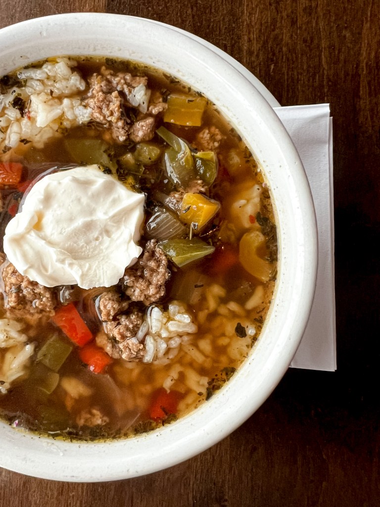 A bowl of stuffed pepper soup with rice, ground meat, and colorful peppers, topped with a white dollop.