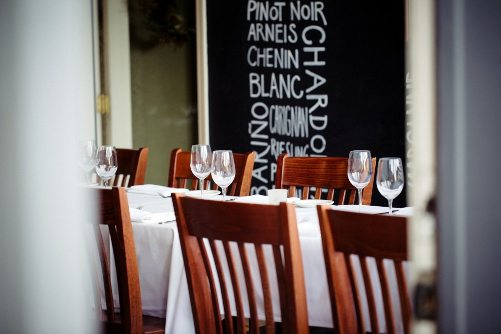Wooden chairs surround tables set with white linens and wine glasses beside a chalkboard listing wine varieties.