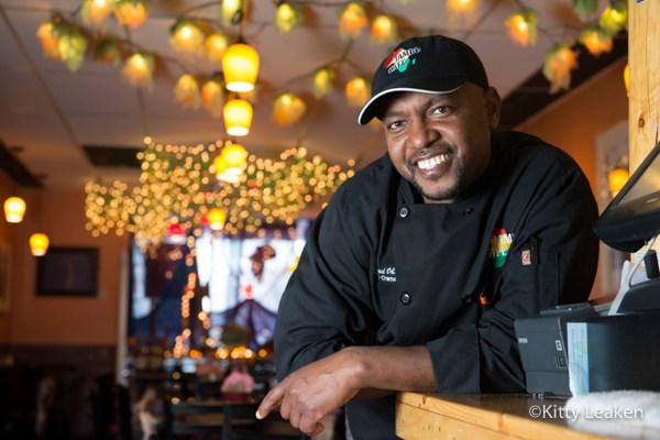 Chef Ahmed Obo smiles while leaning on a wooden counter inside Jambo Cafe, with string lights behind him.