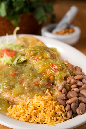 Chile rellenos covered in green chile sauce on a white plate with Spanish rice, pinto beans, shredded lettuce, and tomato.