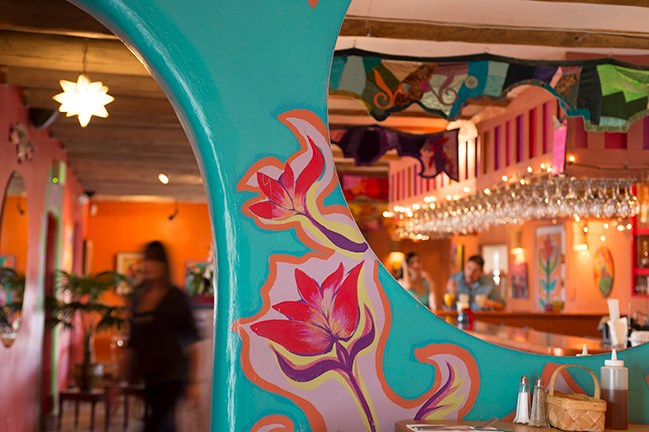 A turquoise arch painted with colorful flowers frames a view of La Choza’s orange-walled bar, hanging glassware, and string lights.