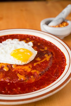 A fried egg with a bright yellow yolk tops a cheese enchilada covered in red chile sauce on a white plate.