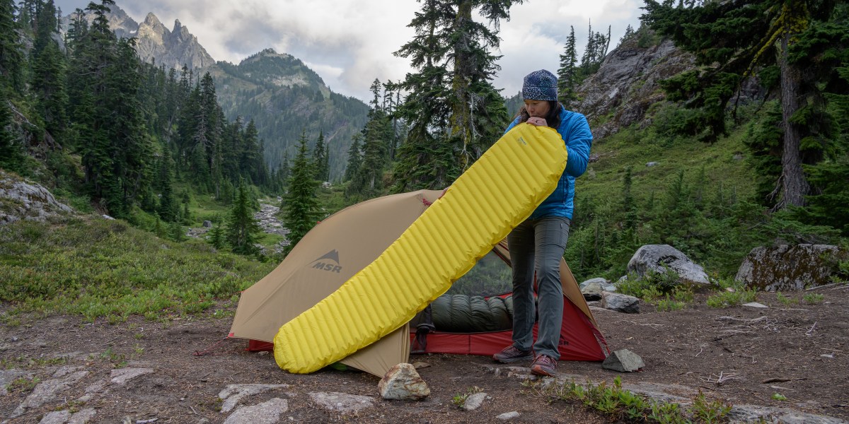 woman blows into sleeping mat to inflate it with a tent, trees, and mountains in background