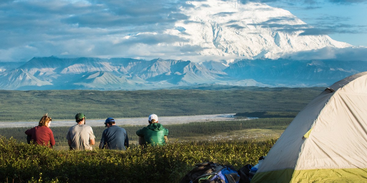 four people sitting near a tent on a hill, looking at a distant, snowy mountain