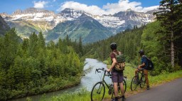 two cyclists stand beside bikes looking out at a river and mountains