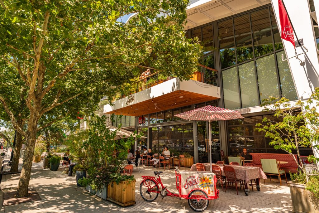 The exterior of a restaurant with greenery, striped umbrellas, and a cart attached to a bike.